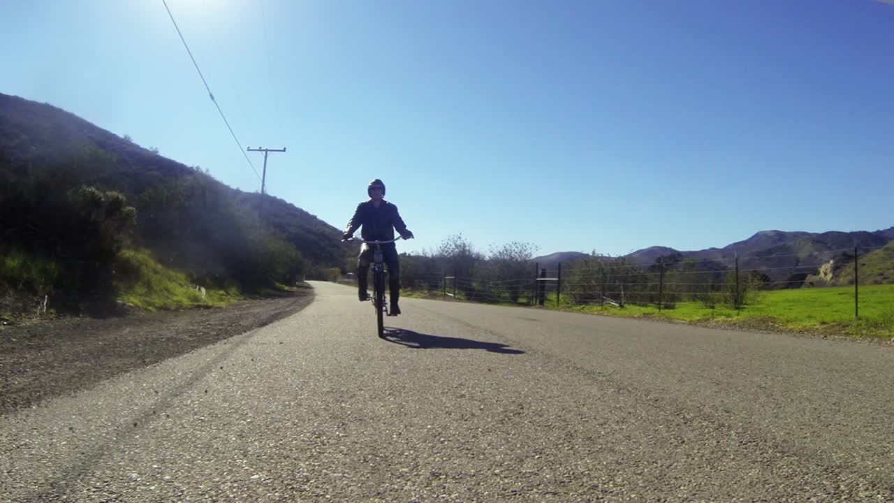 가죽 옷을 입은 남자가 2차선 도로 2에서 시골을 통해 전동 자전거를 탄다
