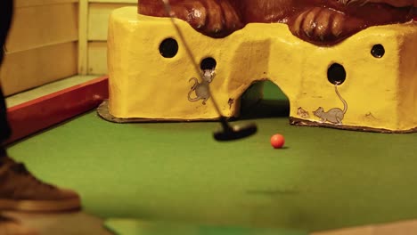 a player putts a red ball through a cheese-shaped obstacle on a mini-golf course.