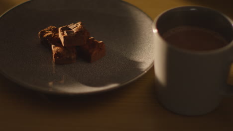 close up de una persona recogiendo una taza de bebida de chocolate caliente humeante en la mesa junto al plato de brownies