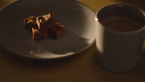 close up of person picking up steaming cup of hot chocolate drink on table next to plate of brownies