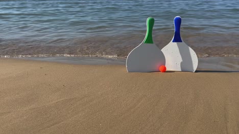 Beach-tennis-paddle-rackets-and-red-balls-on-sandy-beach-with-waves-breaking-on-shore