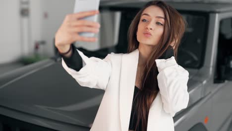 Smiling-girl-in-a-white-jacket-makes-a-photo-on-the-background-of-the-car-in-the-showroom