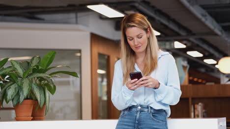 casually dressed young businesswoman checking mobile phone in modern open plan workplace