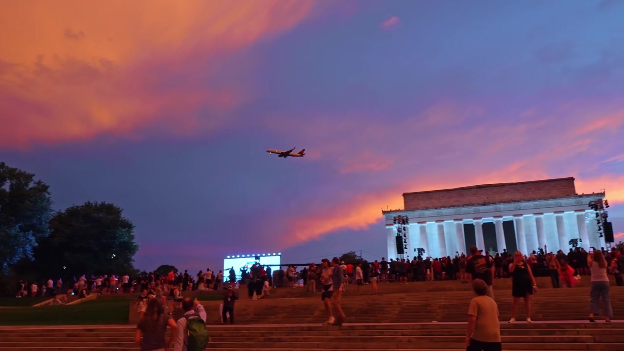 Washington, USA, 13 June 2026 : Airplane over Lincoln Memorial crowd at dusk, event screens, staircases, trees and pink clouds appear.