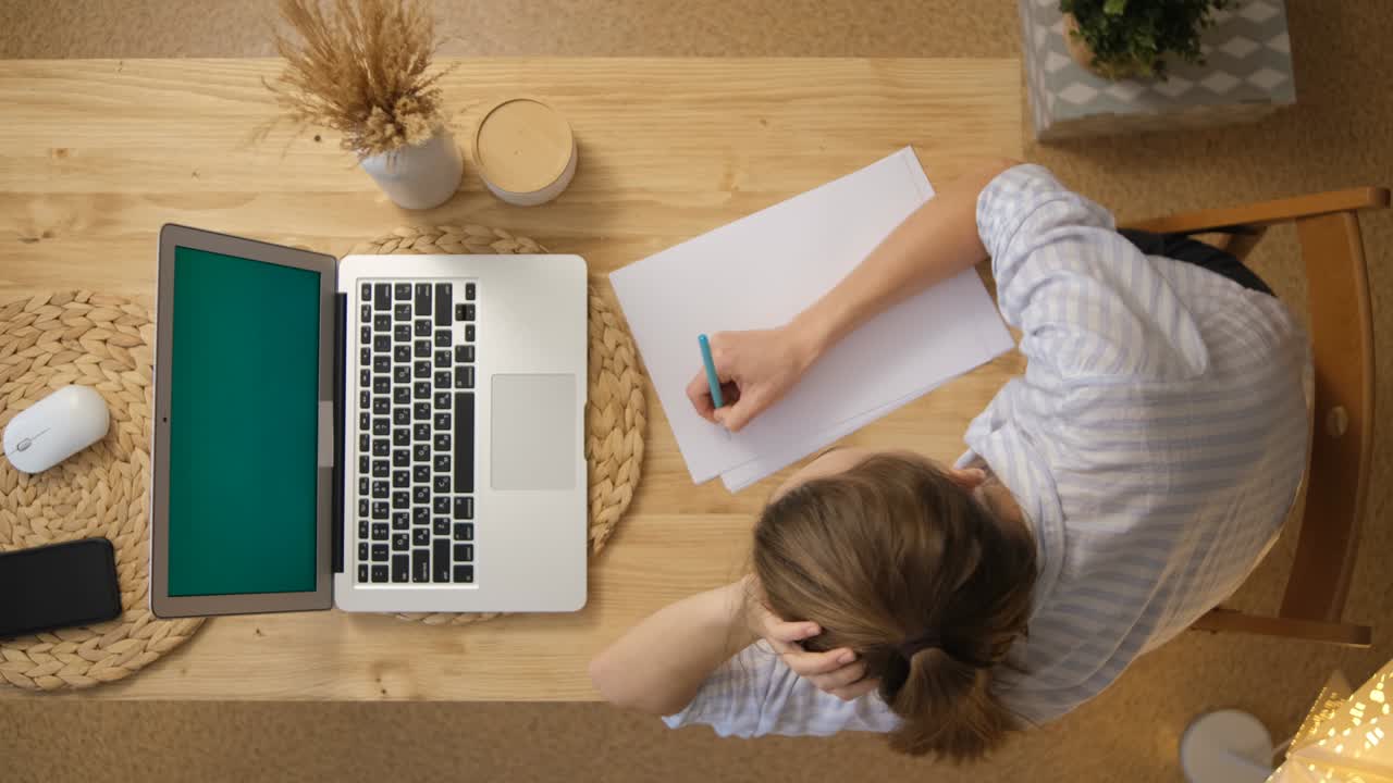 The girl records a lecture from remote training. A laptop with a green screen. Top view. Conference on the Internet. Make important notes on paper.