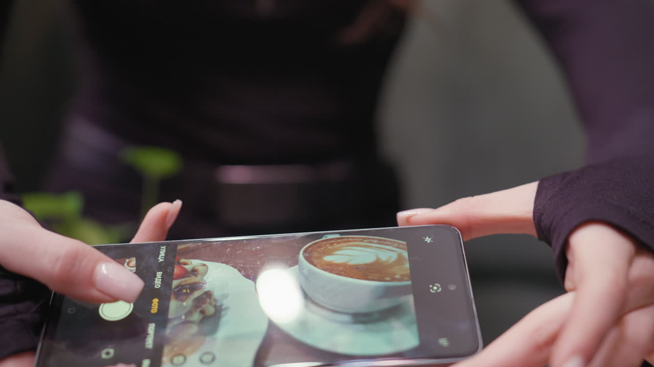 Close view of lady's hand holding phone while capturing top-down photo of latte and waffle. Focus is on manicured fingers and phone screen, with blurred outfit and indoor setting in background