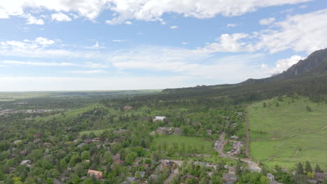 Panorámica-Aérea-A-La-Derecha-Del-Cálido-Sol-Golpeando-Las-Montañas-Flatiron-De-Boulder-Colorado-Sobre-El-Parque-Chautauqua-Con-Pinos-Verdes-Y-Cielos-Azules-Con-Nubes-En-Un-Hermoso-Día-De-Verano-Para-Hacer-Senderismo