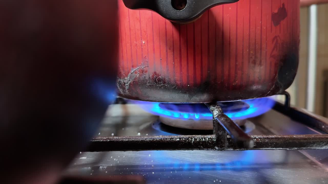 Close-up of a hand lighting a gas stove burner with a lighter and placing a pot on the cooktop, showing blue flame, indoor kitchen action, and home cooking preparation