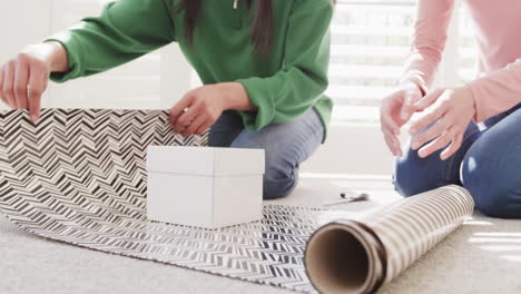 Low section of biracial mother and adult daughter wrapping christmas gift at home, slow motion