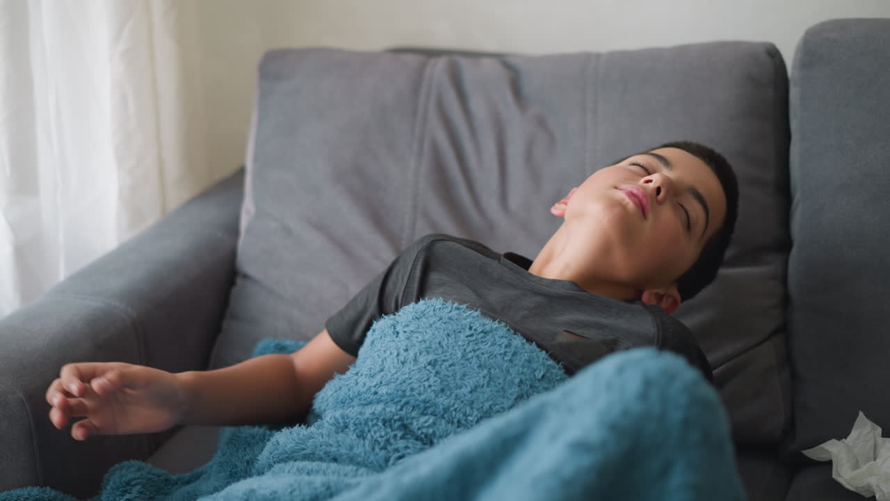 Studente in vacanza si riposa sul divano avvolto in una coperta, si sistema per stare più comodo con un fazzoletto usato accanto, ambiente interno rilassato con luce naturale soffusa proveniente dalla finestra