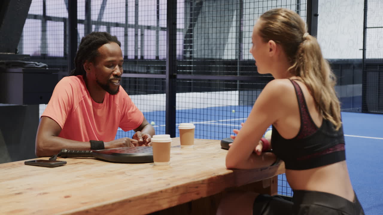 Two diverse athletes enjoying coffee and conversation after padel tennis match on indoor court