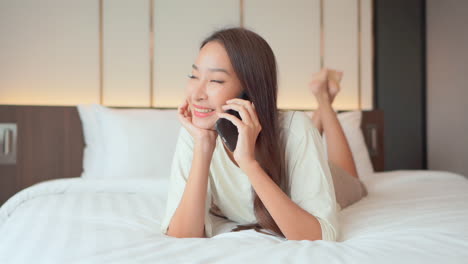 happy and beautiful thai young woman lying on bed talks on mobile phone