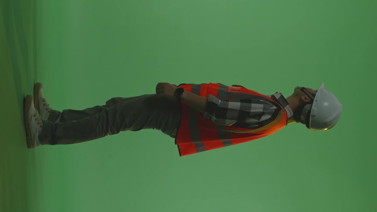 vista lateral completa del cuerpo de un ingeniero masculino asiático con casco de seguridad mirando a su alrededor mientras está de pie en el estudio de fondo de pantalla verde, durante la puesta o el amanecer