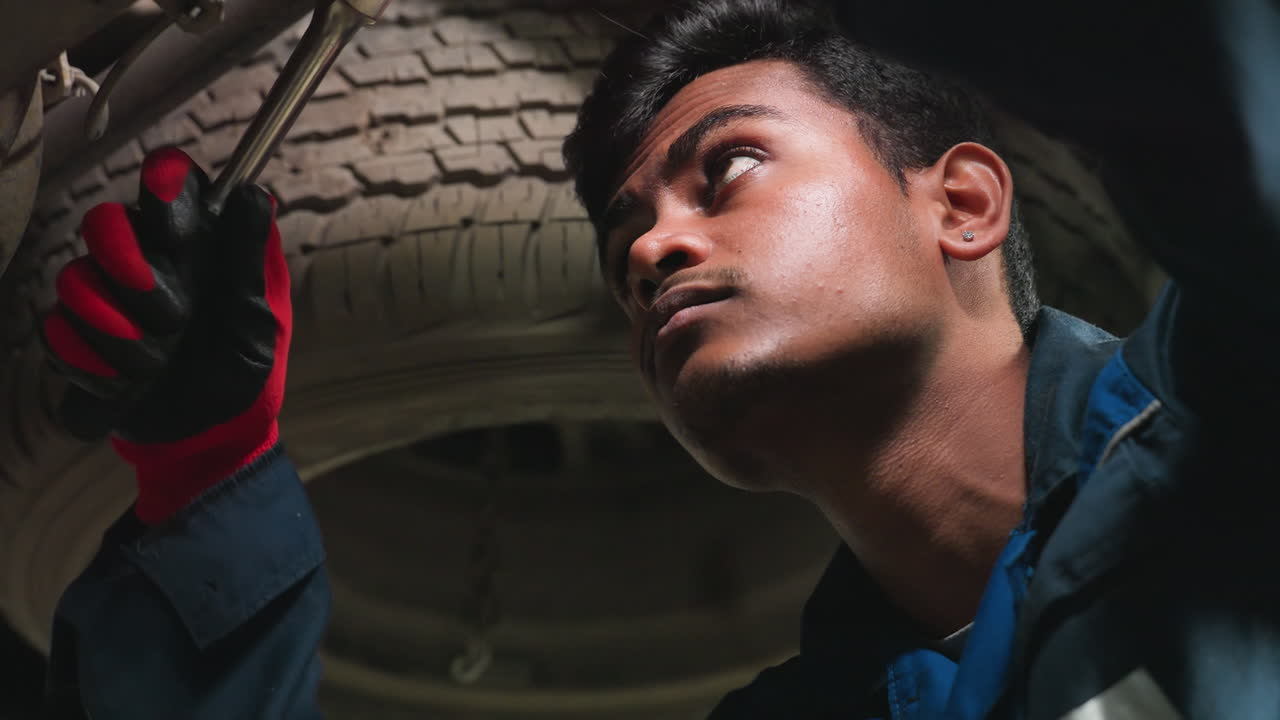 ingeniero indio que trabaja debajo de un coche, apretando el perno con expresión enfocada, la luz se refleja en la cara, usando guantes y uniforme azul, involucrado en la reparación de automóviles, inspeccionando la mecánica de vehículos