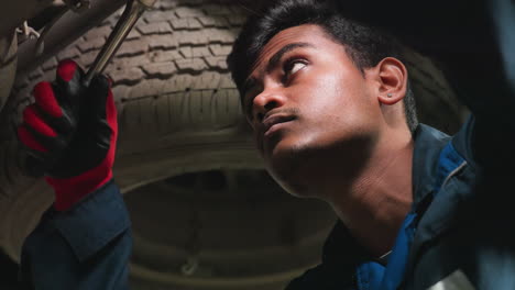 ingeniero indio que trabaja debajo de un coche, apretando el perno con expresión enfocada, la luz se refleja en la cara, usando guantes y uniforme azul, involucrado en la reparación de automóviles, inspeccionando la mecánica de vehículos