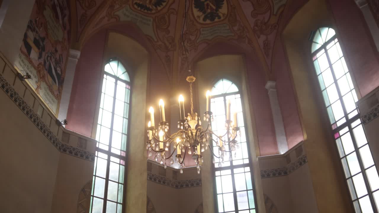 Elegant chandelier illuminating a historic church interior with tall stained glass windows and decorative wall art.