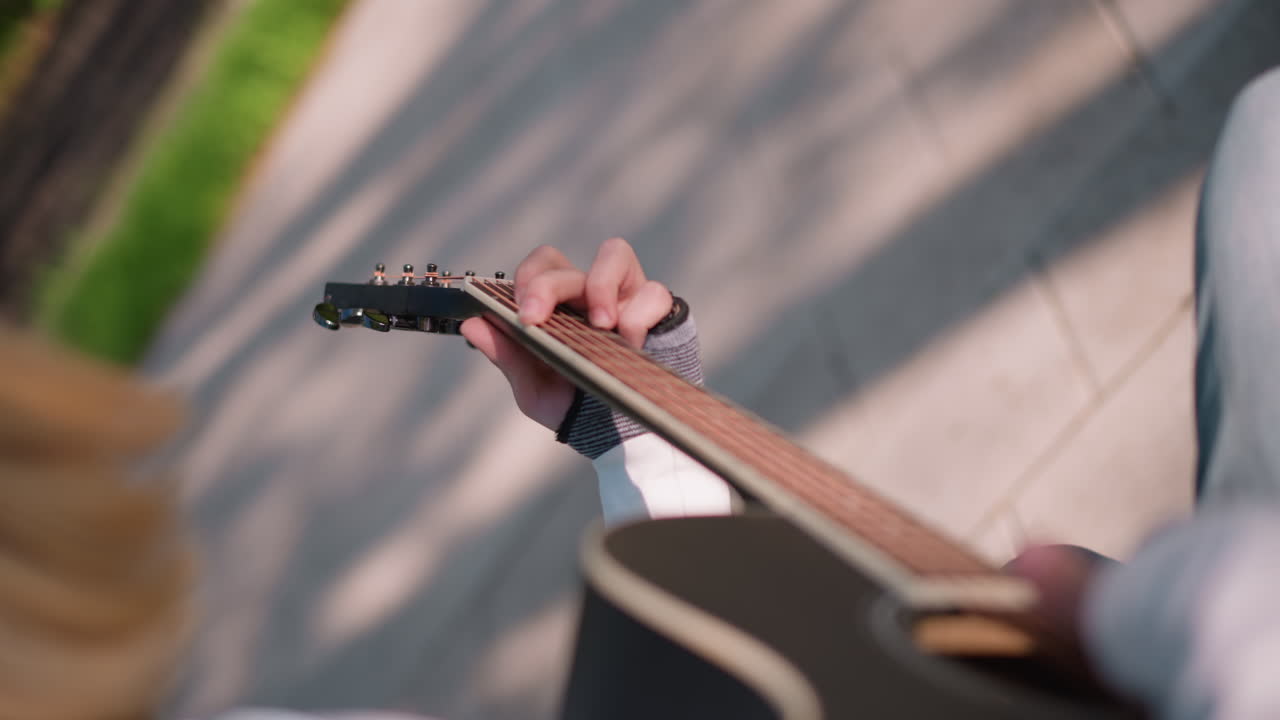 overhead closeup guitarist playing acoustic, focus on fretting hand, denim jeans, blonde hair fringe, sunlight stripe, bench setting, upbeat fingerpicking tutorial, busking mood, subtle shadows, warm