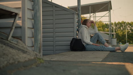 girl in jeans seated on ground with thoughtful expression, resting hand on head, black bag nearby, with trees in the distant background