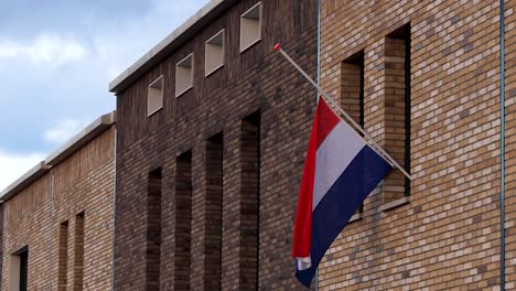 dutch national flag hanging half-mast on the exterior facade of a modern building in commemoration of those fallen in war times against an overcast blue sky in the background