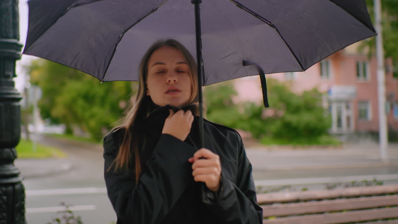 Female with long hair wearing black coat standing under umbrella on city sidewalk near bench during cold and windy overcast day looking sideways with thoughtful expression