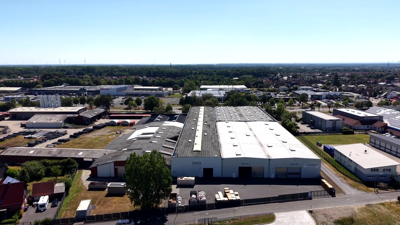 Stor hvidtaget lagerbygning ligger centralt i industripark i Tyskland, spredte erhvervsbygninger og rækkehustage strækker sig mod skovbrynet under klar sommerhimmel. Droneoverflyvning
