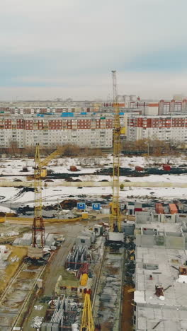 giant construction cranes on building in progress against distant town with houses and infrastructure aerial view