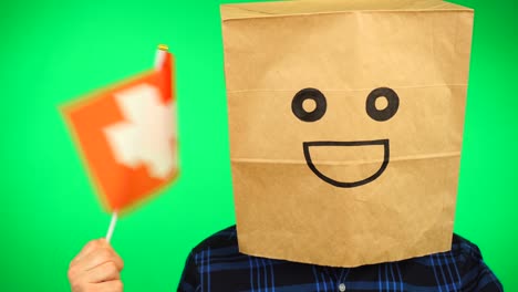 portrait of man with paper bag on head waving swiss flag with smiling face against green background.