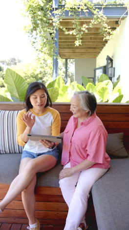 vertical video: using tablet, young woman showing something to senior woman, sitting on bench