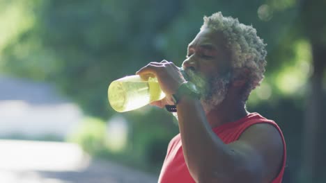 Afroamerikanischer-älterer-Mann,-Der-Draußen-Trainiert-Und-Auf-Der-Straße-Aus-Einer-Wasserflasche-Trinkt
