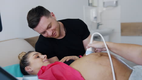 caucasian male smiles to his wife undergoing ultrasonic pregnancy check up. doctor quickly moves ultrasonic device by the big belly covered with gel.