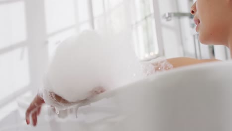 video of smiling biracial woman lying in bathtub in bubble bath in bathroom