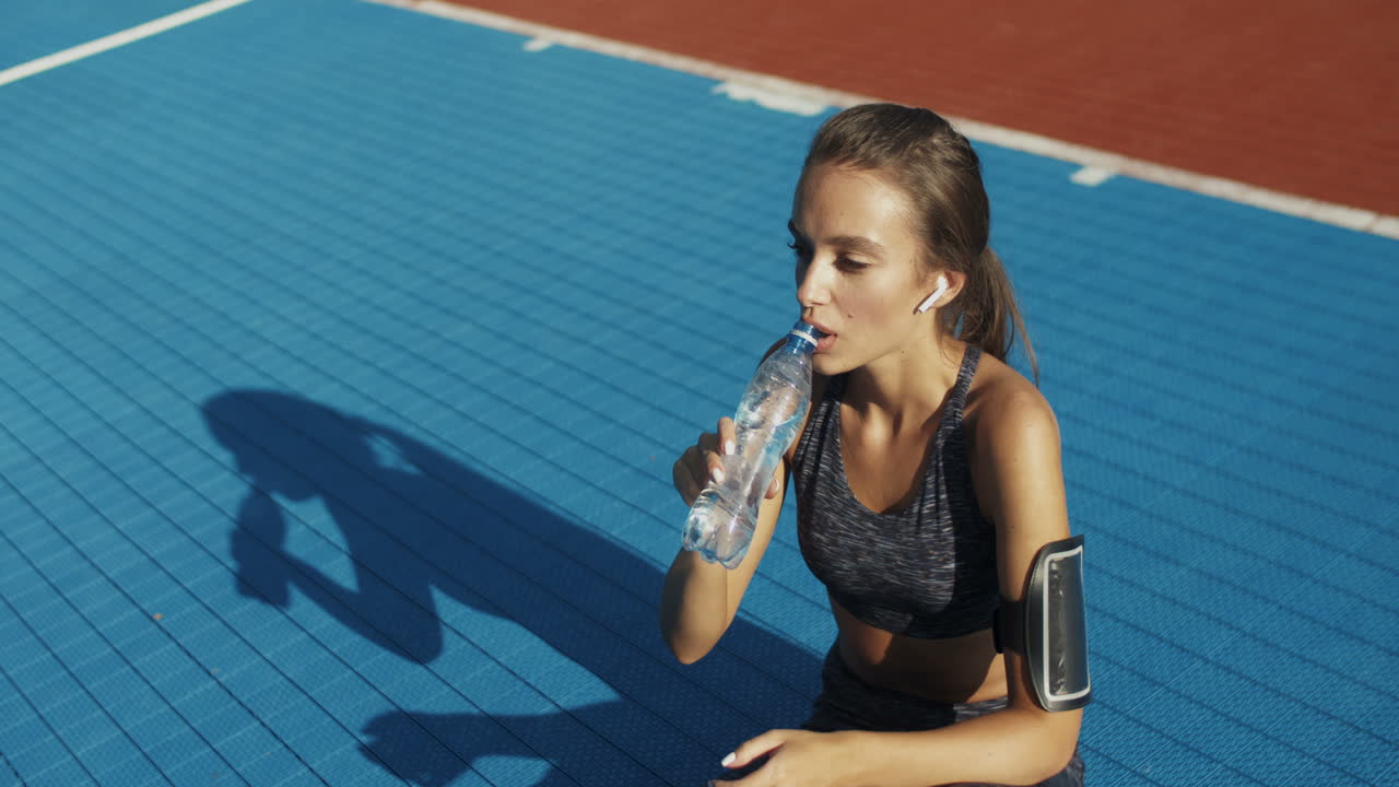 mujer fitness sentada en la cancha deportiva y bebiendo agua fría después del entrenamiento en un día de verano 1