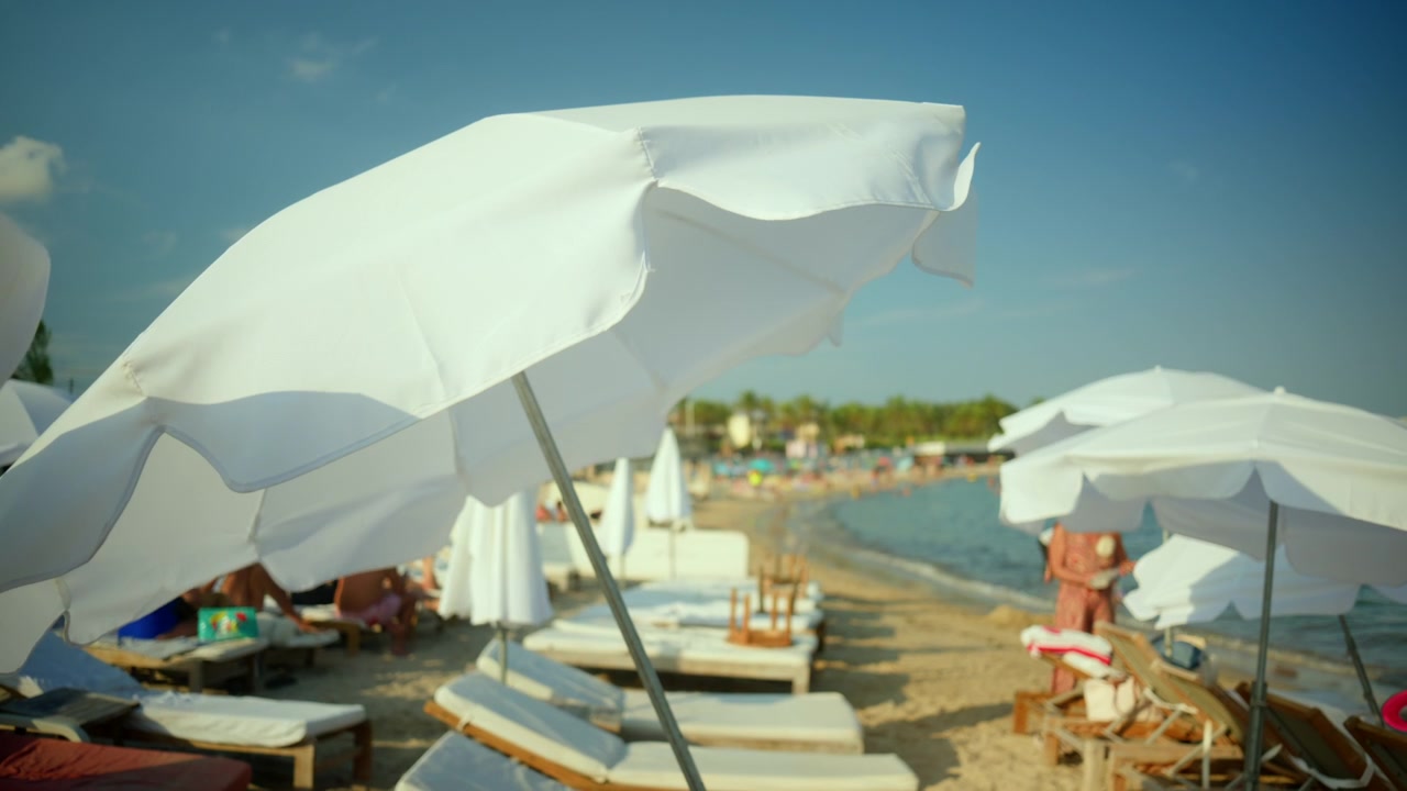Ombrelloni da spiaggia bianchi che offrono ombra su una spiaggia sabbiosa con persone che si rilassano e nuotano in mare in una giornata di sole.