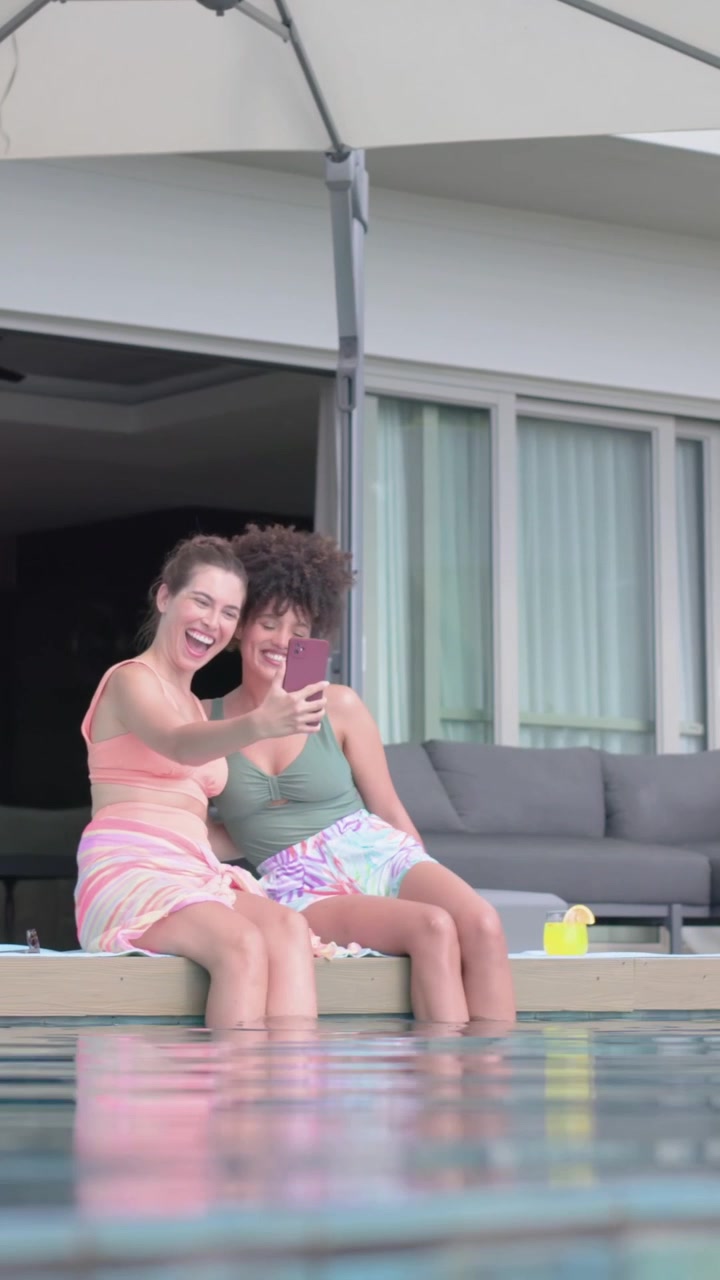 Two women tapping phone for social pool selfies, hearts floating over phone, raising yellow cup