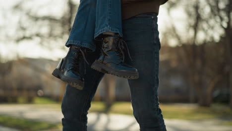 a man wearing a brown jacket lifts a girl who is wearing blue jeans and black boots. the scene captures an outdoor moment with sunlight filtering through trees in the background