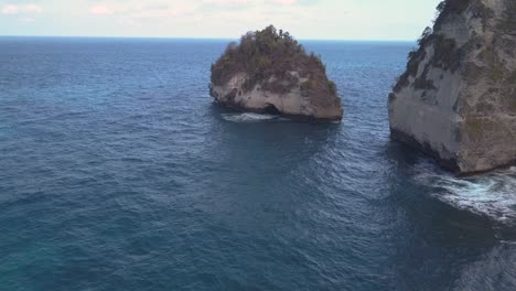 Aerial-view-of-the-crystal-blue-water-of-diamond-beach-in-Nusa-Penida,-Indonesia-with-a-prominent-large-cliff-in-the-foreground