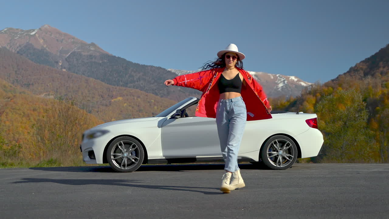 가을의 산에서 색 컨버터블 자동차에 의해 춤을 추는 여자