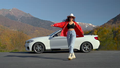 mujer bailando por un coche descapotable blanco en las montañas de otoño