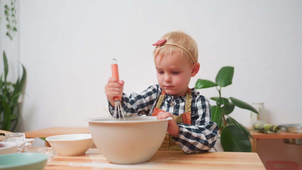 Un bambino mescola la farina con una frusta in una ciotola grande durante la preparazione della cottura, indossa un grembiule e si concentra intensamente, circondato da ciotole e utensili da cucina mentre impara le basi della cucina