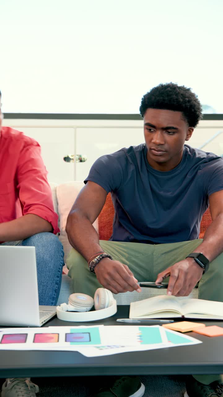 Vertical video: Diverse male coworkers on couch reviewing laptop screen and writing planning notes