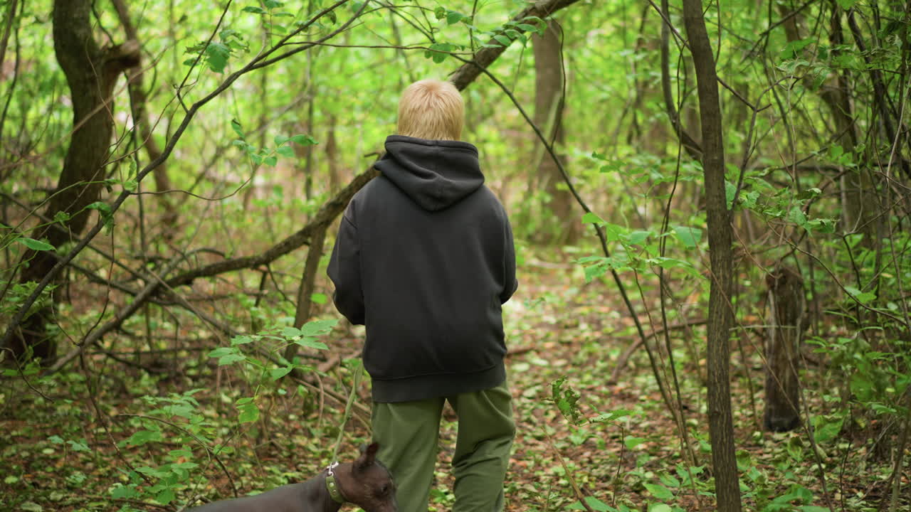 White Boy Walking Through Dense Woods With Loyal Dog At Side, Hooded Child Explorer Threading Narrow Trail, Quiet Discovery And Companionship Among Green Undergrowth And Tangled Branches