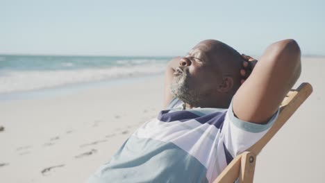 hombre afroamericano mayor feliz sentado en una silla de sol en la playa, en cámara lenta