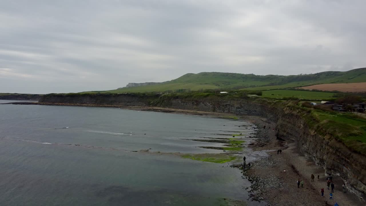 Drone video around Kimmeridge Bay, Dorset on a cloudy day. Gentle waves in the bay as the footage moves over the rocks and pebble beach and the turns gently to the left around the rest of the bay.