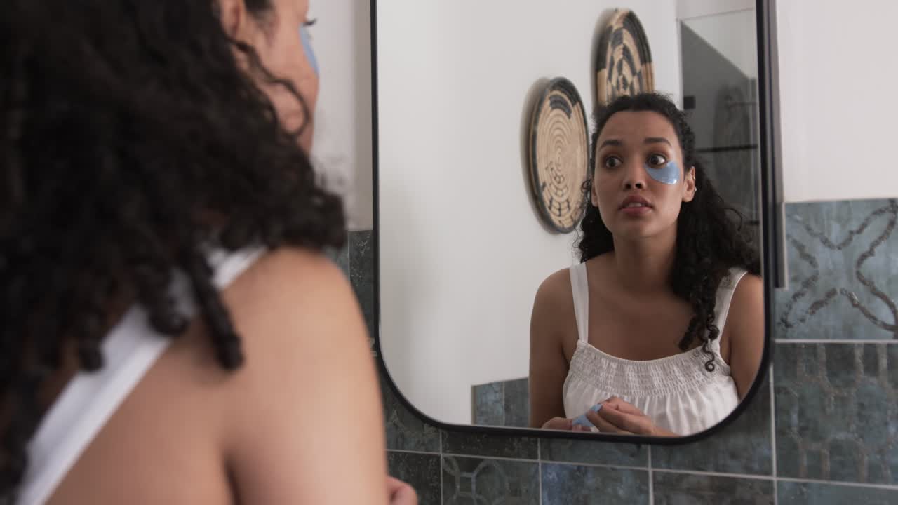Donna che controlla la pelle, applica e rimuove patch blu sotto gli occhi davanti allo specchio del bagno per ridurre il gonfiore.