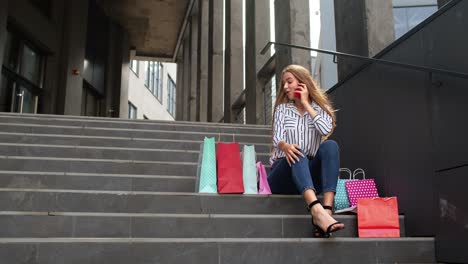 girl sitting on stairs with bags talking on mobile phone about sale in shopping mall in black friday