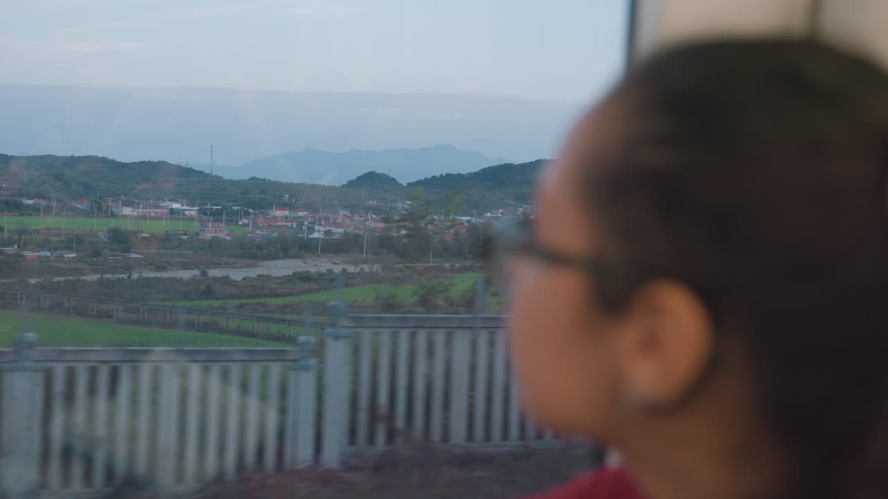 Girl looking through train window at passing countryside during dusk, blurred reflections on glass, contemplative mood, seated inside carriage with glasses, distant hills and village visible