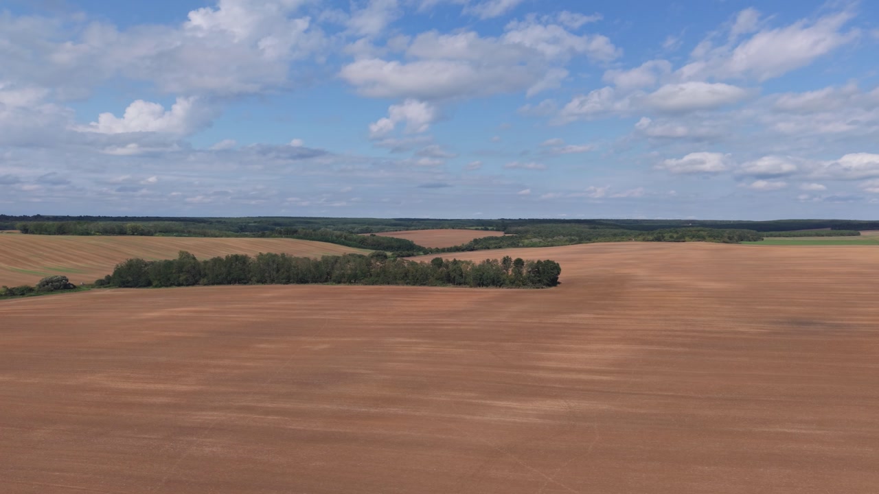 An aerial view of wide open farmland with freshly plowed fields under a partly cloudy sky. The landscape features gentle rolling hills and tree lines stretching toward the horizon.
