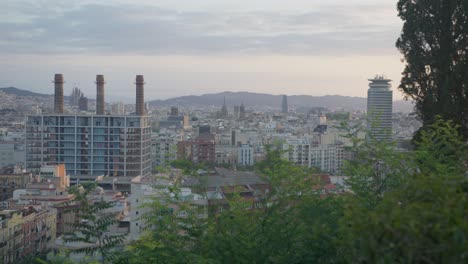 Vista-De-Alta-Resolución-A-Través-De-árboles-Pintorescos,-Vista-De-Fábrica,-Horizonte-De-La-Ciudad-De-Barcelona,-España,-Con-Amanecer-Dorado-En-6k-Mientras-Los-Pájaros-Vuelan