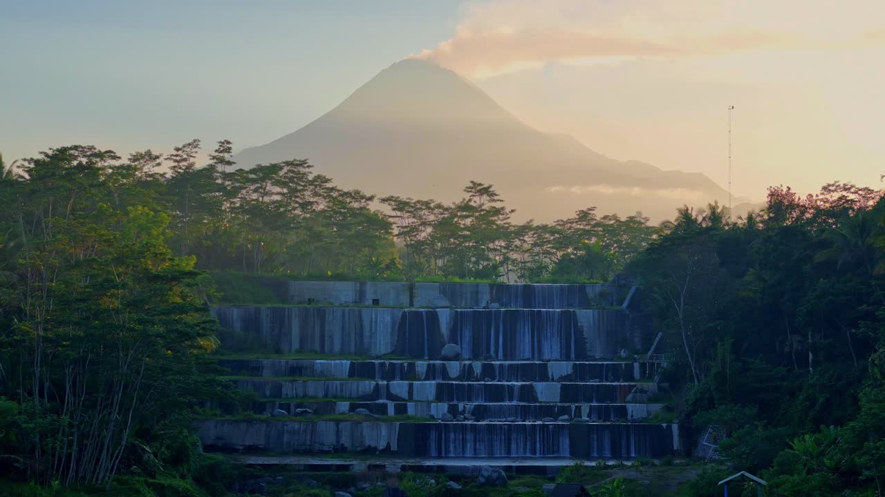 Reveal drone shot of terraced waterfall on the middle of forest with volcano emitting smoke and sunrise sky on the background. Watu Purbo waterfall and Merapi volcano. 4k Aerial view.