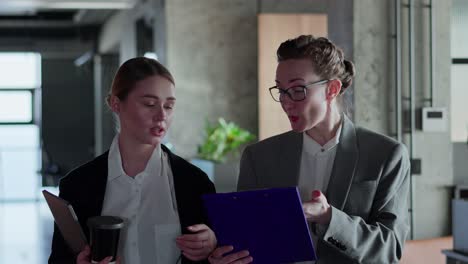 two businesswoman girls in business clothes and jackets walk along the corridor in a modern office and communicate about specific paper on a tablet